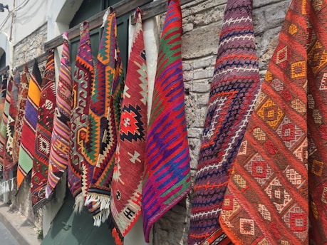 Several colorful and intricately patterned rugs hang side by side against a stone wall. The patterns include geometric shapes and are woven in vibrant colors such as red, orange, purple, blue, and green.