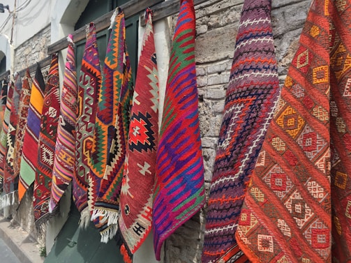 Several colorful and intricately patterned rugs hang side by side against a stone wall. The patterns include geometric shapes and are woven in vibrant colors such as red, orange, purple, blue, and green.