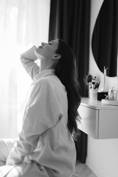 A woman with long hair is sitting in profile, looking upward with a contemplative expression. She is wearing a loose-fitting white shirt and appears to be next to a vanity with beauty products and a round mirror. The scene is softly lit, with curtains in the background creating a gentle ambiance.