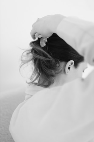 A black and white photograph captures a person with long hair being gathered and styled. The focus is on the back of the head and shoulders, with a hand visible holding the hair.