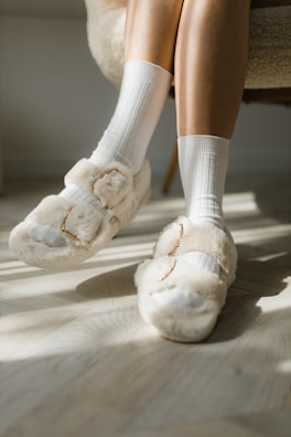 A pair of legs wearing white socks and fluffy beige slippers resting on a light wooden floor. Soft sunlight casts shadows across the floor, creating a warm and cozy atmosphere.