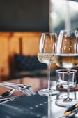 Two wine glasses filled with a sparkling beverage are placed on a reflective table surface. Silverware is neatly arranged beside the glasses. The background appears softly blurred, suggesting an elegant and serene dining setting.