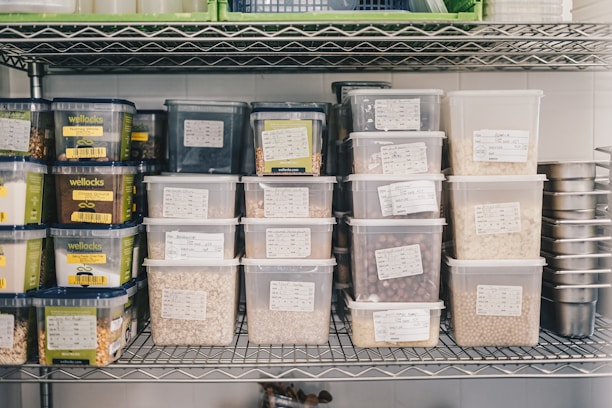 A metal shelf is neatly organized with various labeled plastic containers filled with grains, nuts, and seeds. The labels on the containers suggest careful categorization of contents, with some containers branded as 'wellocks.' The surrounding environment appears to be a storage or kitchen area, with additional metal containers stacked on the right.