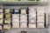 A metal shelf is neatly organized with various labeled plastic containers filled with grains, nuts, and seeds. The labels on the containers suggest careful categorization of contents, with some containers branded as 'wellocks.' The surrounding environment appears to be a storage or kitchen area, with additional metal containers stacked on the right.