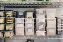 A metal shelf is neatly organized with various labeled plastic containers filled with grains, nuts, and seeds. The labels on the containers suggest careful categorization of contents, with some containers branded as 'wellocks.' The surrounding environment appears to be a storage or kitchen area, with additional metal containers stacked on the right.
