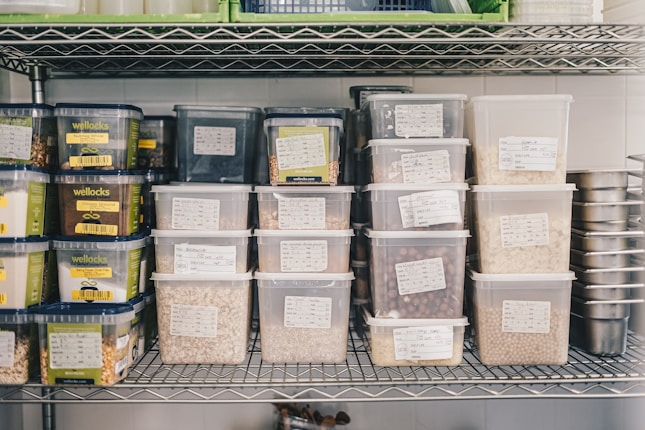 A metal shelf is neatly organized with various labeled plastic containers filled with grains, nuts, and seeds. The labels on the containers suggest careful categorization of contents, with some containers branded as 'wellocks.' The surrounding environment appears to be a storage or kitchen area, with additional metal containers stacked on the right.