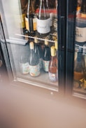 Several wine bottles are displayed inside a glass-doored refrigerator. The bottles have colorful labels and are positioned on metal shelves. The fridge also has partially obscured areas with condensation on the glass.