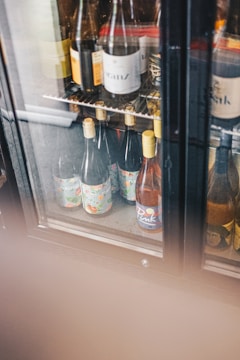 Several wine bottles are displayed inside a glass-doored refrigerator. The bottles have colorful labels and are positioned on metal shelves. The fridge also has partially obscured areas with condensation on the glass.