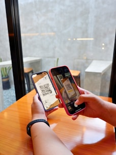 A person is holding two smartphones, one in each hand, on a wooden table. One of the phones has a QR code on its screen, while the other phone is capturing or scanning the QR code with its camera application. The scene is set indoors, by a glass window, with some stools and potted plants visible in the background. The person is wearing a dark wristband on their left wrist.