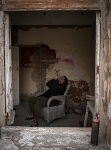 A person sitting on a wicker chair in a dilapidated room, covering their face with their hands. The walls are peeling, with visible patches of different colors and textures. Debris is scattered on the floor, and there are remnants of construction or destruction, including pipes and a partial door frame. An aerosol can stands on the ground in the foreground.