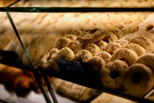 A variety of pastries are displayed behind a glass counter, arranged in neat rows. The pastries appear to be golden brown with a dusting of sugar, and a small sign indicates the price per kilogram.