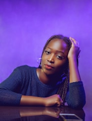 A person with braided hair is leaning on a reflective surface, with one hand resting on their head. They are wearing a dark blue textured top and hoop earrings. The background is a gradient of purple hues, creating a vibrant and stylish atmosphere.