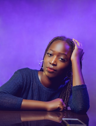 A person with braided hair is leaning on a reflective surface, with one hand resting on their head. They are wearing a dark blue textured top and hoop earrings. The background is a gradient of purple hues, creating a vibrant and stylish atmosphere.