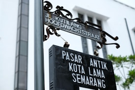 A metal street sign with ornate detailing at the top and the words 'Jl. Kutlang Hoofdwachtstraat' displayed, hanging above a black rectangular sign that reads 'Pasar Antik Kota Lama Semarang'. The background features the facade of a white building with tall windows and some greenery visible.