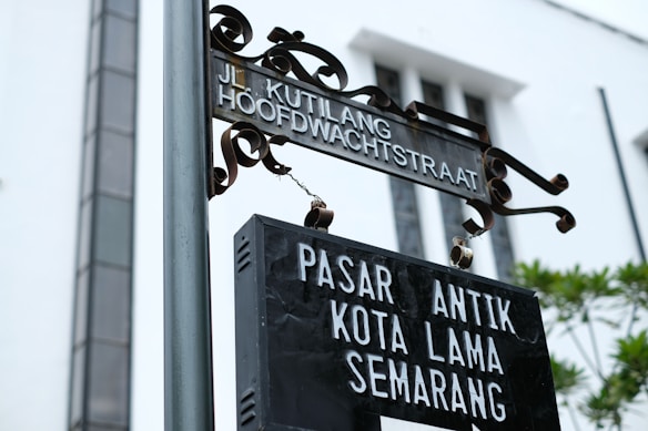 A metal street sign with ornate detailing at the top and the words 'Jl. Kutlang Hoofdwachtstraat' displayed, hanging above a black rectangular sign that reads 'Pasar Antik Kota Lama Semarang'. The background features the facade of a white building with tall windows and some greenery visible.