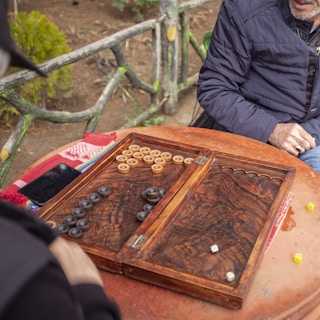 Two people are sitting at a round table outdoors, playing a board game with wooden pieces and dice. The board has a rich, dark wood texture, and there is a smartphone lying on the table. The background includes greenery and a rustic fence.