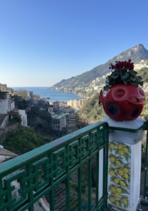 A scenic view of a coastal town with verdant hills and a clear blue sky. In the foreground, a decorative element adorned with lemons and a vibrant red ceramic object with plants is placed on a green railing. The background features a coastline with a calm sea and scattered buildings.