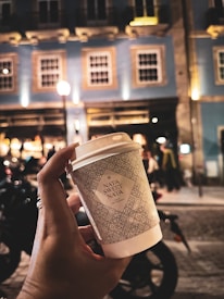 A person's hand is holding a takeaway paper cup against a backdrop of a well-lit, bustling street with a blue building featuring multiple windows. The street appears lively with blurred images of people and vehicles, possibly at night. The cup has intricate patterns and the words 'Nata Sweet Nata' printed on it.