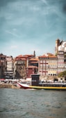 A scenic riverside view features a vibrant cityscape with colorful historic buildings. The foreground has a boat on the water and people walking along the promenade, capturing a lively atmosphere under a clear sky.