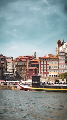 A scenic riverside view features a vibrant cityscape with colorful historic buildings. The foreground has a boat on the water and people walking along the promenade, capturing a lively atmosphere under a clear sky.