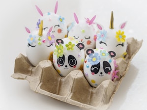 Eggs placed in a cardboard carton are decorated with designs resembling panda faces and unicorns. Each egg is adorned with colorful floral stickers, and some feature drawn-on eyes and noses to create whimsical expressions.