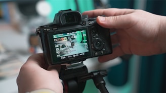 A person is holding a DSLR camera, reviewing an image on its screen. The camera is mounted on a tripod and appears to be pointed at a cluttered desk or workspace, which is reflected on the camera display.
