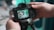 A person is holding a DSLR camera, reviewing an image on its screen. The camera is mounted on a tripod and appears to be pointed at a cluttered desk or workspace, which is reflected on the camera display.