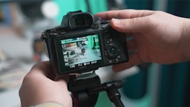 A person is holding a DSLR camera, reviewing an image on its screen. The camera is mounted on a tripod and appears to be pointed at a cluttered desk or workspace, which is reflected on the camera display.