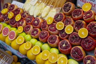 A colorful display of fresh fruit at a market featuring sliced pomegranates, oranges, grapefruits, and whole apples. The fruits are neatly arranged and vibrant in their colors, with some wrapped pineapple pieces visible in the background.