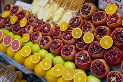 A colorful display of fresh fruit at a market featuring sliced pomegranates, oranges, grapefruits, and whole apples. The fruits are neatly arranged and vibrant in their colors, with some wrapped pineapple pieces visible in the background.