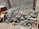 A pile of mixed scrap metal and machinery parts is stacked against a textured concrete wall. Various components, including pipes, rods, and mechanical elements, are visible, creating a chaotic arrangement. The scene is in an outdoor setting with dirt and debris scattered on the ground.