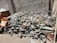 A pile of mixed scrap metal and machinery parts is stacked against a textured concrete wall. Various components, including pipes, rods, and mechanical elements, are visible, creating a chaotic arrangement. The scene is in an outdoor setting with dirt and debris scattered on the ground.