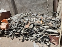 A pile of mixed scrap metal and machinery parts is stacked against a textured concrete wall. Various components, including pipes, rods, and mechanical elements, are visible, creating a chaotic arrangement. The scene is in an outdoor setting with dirt and debris scattered on the ground.
