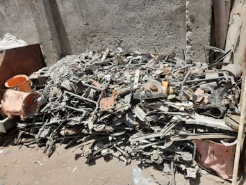A pile of mixed scrap metal and machinery parts is stacked against a textured concrete wall. Various components, including pipes, rods, and mechanical elements, are visible, creating a chaotic arrangement. The scene is in an outdoor setting with dirt and debris scattered on the ground.