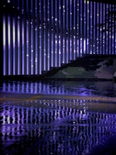 Vertical light bars create a striking pattern against a backdrop, reflecting vividly on a wet surface. The ambiance is enhanced by the mix of artificial and natural elements, including a graffiti-style drawing along one side.