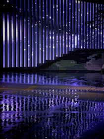 Vertical light bars create a striking pattern against a backdrop, reflecting vividly on a wet surface. The ambiance is enhanced by the mix of artificial and natural elements, including a graffiti-style drawing along one side.