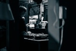 A person wearing headphones is operating a DJ mixer in a dimly lit indoor setting. The background contains blurred elements such as plants and decor, contributing to an intimate and cozy atmosphere.