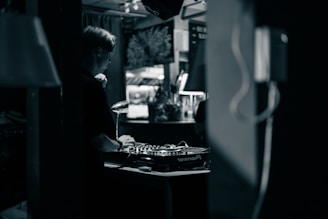 A person wearing headphones is operating a DJ mixer in a dimly lit indoor setting. The background contains blurred elements such as plants and decor, contributing to an intimate and cozy atmosphere.