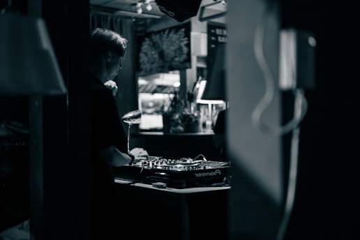 A person wearing headphones is operating a DJ mixer in a dimly lit indoor setting. The background contains blurred elements such as plants and decor, contributing to an intimate and cozy atmosphere.