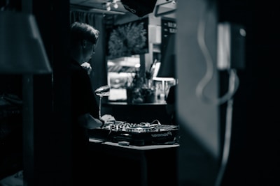 A person wearing headphones is operating a DJ mixer in a dimly lit indoor setting. The background contains blurred elements such as plants and decor, contributing to an intimate and cozy atmosphere.