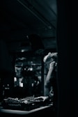 A DJ is focused on mixing music at a dimly lit venue. The setting features a mixing console with various knobs and controls, while the background is blurred, suggesting a lively atmosphere with possible reflections or movement.