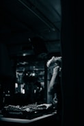 A DJ is focused on mixing music at a dimly lit venue. The setting features a mixing console with various knobs and controls, while the background is blurred, suggesting a lively atmosphere with possible reflections or movement.