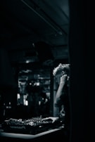 A DJ is focused on mixing music at a dimly lit venue. The setting features a mixing console with various knobs and controls, while the background is blurred, suggesting a lively atmosphere with possible reflections or movement.