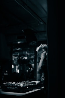 A DJ is focused on mixing music at a dimly lit venue. The setting features a mixing console with various knobs and controls, while the background is blurred, suggesting a lively atmosphere with possible reflections or movement.