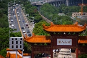 A traditional Chinese gate with a vibrant orange roof stands prominently in the foreground, surrounded by lush green trees. Behind the gate, a busy road filled with vehicles extends into the distance. An elevated railway track runs parallel to the road, while a large bridge looms in the background.