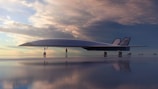 A futuristic aircraft is parked on a reflective surface with a dramatic sky in the background. The plane is sleek in design with the word 'Halcyon' visible on its side. A person stands near the aircraft, highlighting its large size.