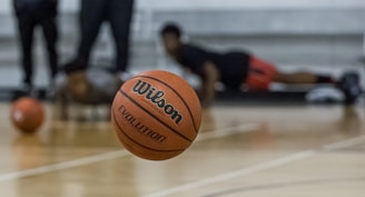 A basketball is prominently in focus mid-air on a court. In the background, individuals are seen in athletic positions, likely involved in training or a workout. The floor is wooden, typical of an indoor basketball court.