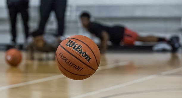 A basketball is prominently in focus mid-air on a court. In the background, individuals are seen in athletic positions, likely involved in training or a workout. The floor is wooden, typical of an indoor basketball court.