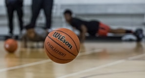 A basketball is prominently in focus mid-air on a court. In the background, individuals are seen in athletic positions, likely involved in training or a workout. The floor is wooden, typical of an indoor basketball court.