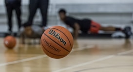 A basketball is prominently in focus mid-air on a court. In the background, individuals are seen in athletic positions, likely involved in training or a workout. The floor is wooden, typical of an indoor basketball court.